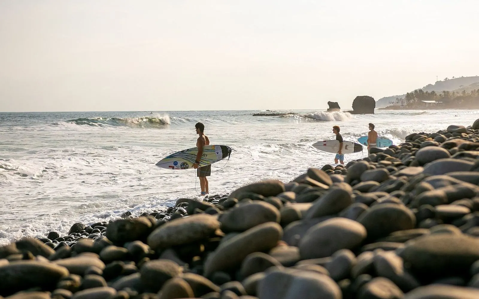 Surfers with boards on a pebble shore beside the sea rocks at El Tunco, El Salvador